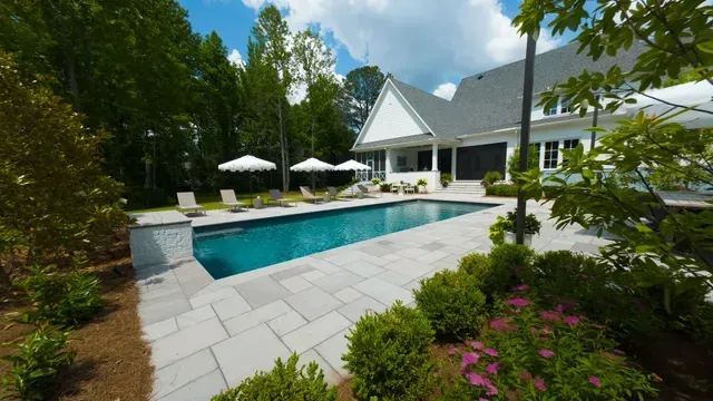 Pool and patio area next to a white house with umbrellas and lounge chairs; trees surround the space.