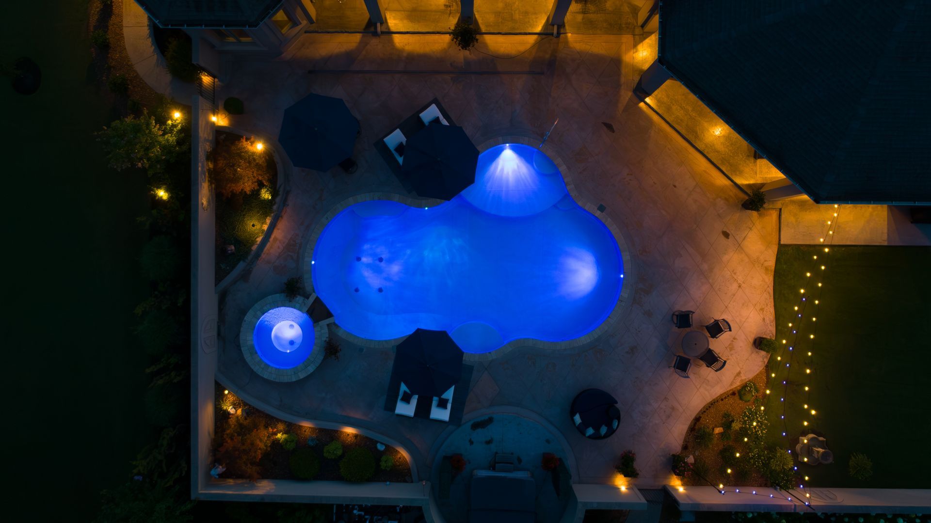 Nighttime aerial view of a glowing blue pool, hot tub, and patio with string lights.