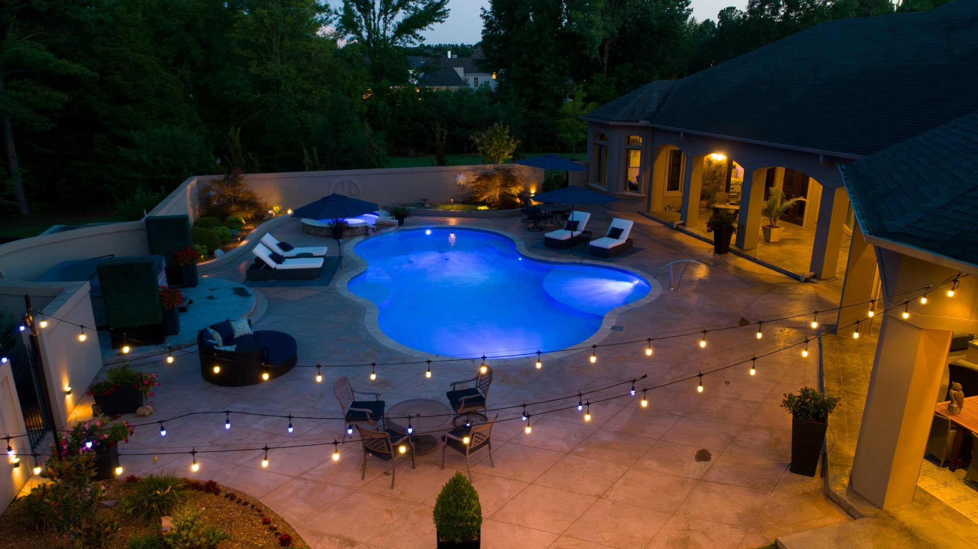 Nighttime backyard with illuminated pool, string lights, and patio furniture.