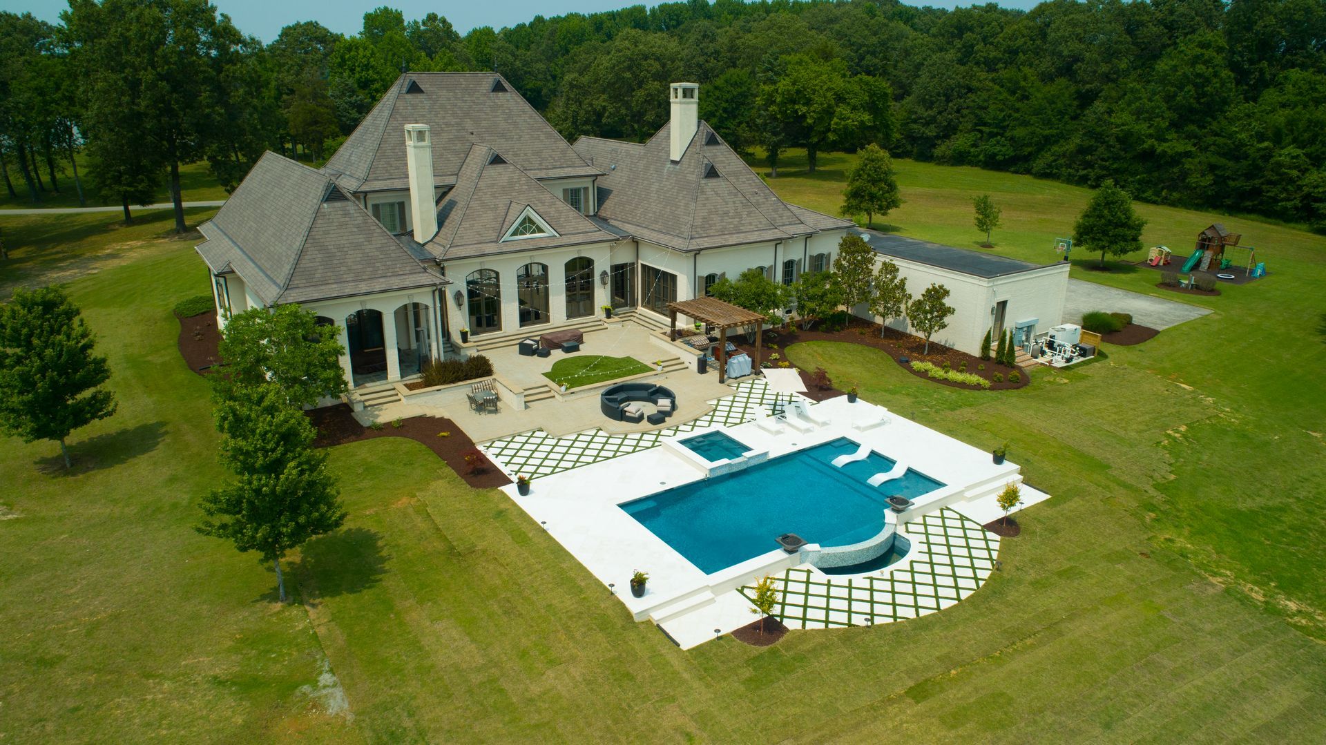 Large white house with pool and patio, green lawn, surrounded by trees, on a sunny day.