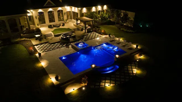 Nighttime view of a luxurious backyard with illuminated pool, hot tub, patio, and house.