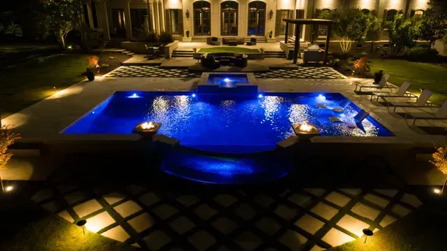 Night view of a luxurious backyard pool with blue water, lit up with surrounding lights, and a house in the background.