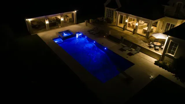 Nighttime aerial view of a luxury home with a blue-lit rectangular pool, patio, and outdoor dining area.
