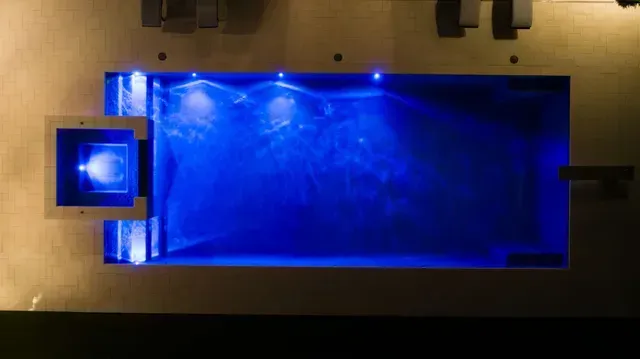 Blue-lit rectangular pool with a square structure on the left side, viewed from above.