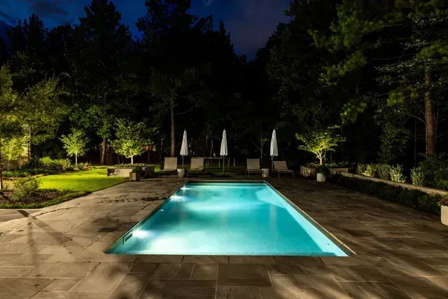 A rectangular pool at night, with trees, lounge chairs, and umbrellas.