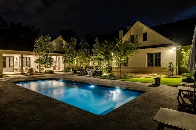 Night view of a house with a lit pool and patio. Trees and building highlighted by outdoor lights.