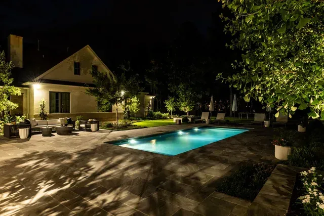 Nighttime view of a lit swimming pool beside a house with outdoor seating, trees and landscaping.