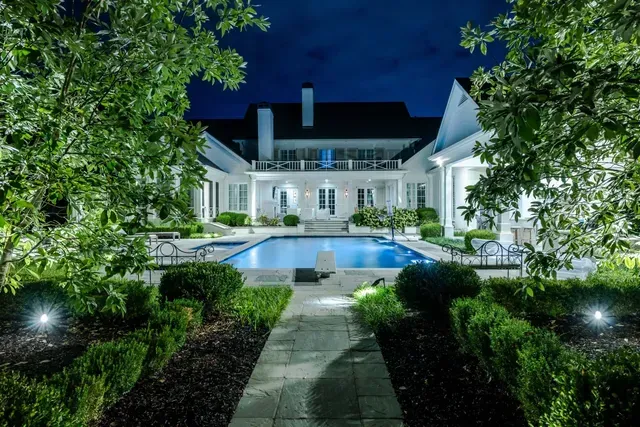Night view of a white mansion with pool, lit by landscape lighting, framed by greenery and a dark blue sky.