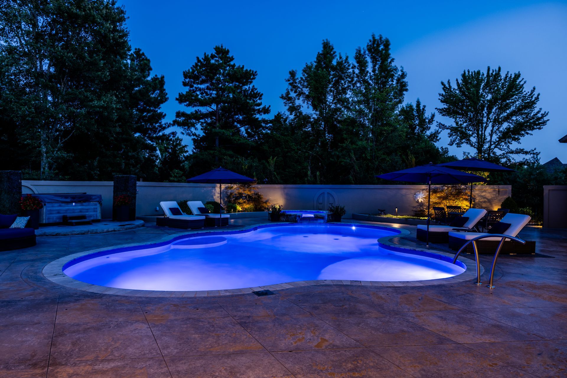 Nighttime backyard pool with blue lights, lounge chairs, and umbrellas.