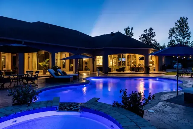 Night view of a luxurious home with a blue-lit pool and patio furniture, with a dark blue sky.