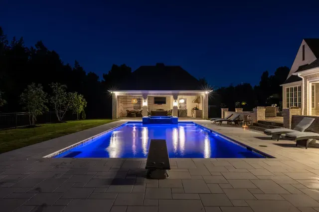 Nighttime view of a lit blue pool with diving board, adjacent to a covered patio and a house.