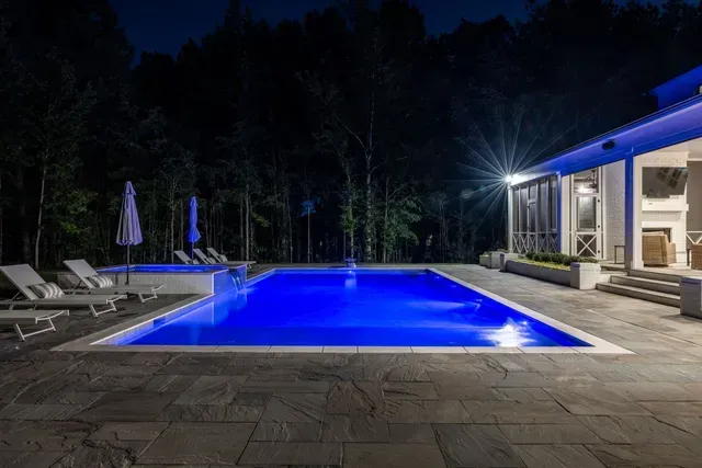 Night view of a blue-lit swimming pool next to a house with trees in the background.