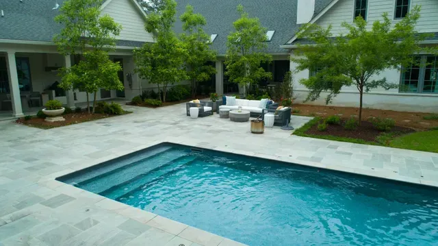 A rectangular pool in a paved courtyard with seating area and a white house in the background.
