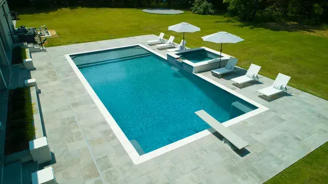 Outdoor pool with diving board, hot tub, lounge chairs, and umbrellas on a sunny day.