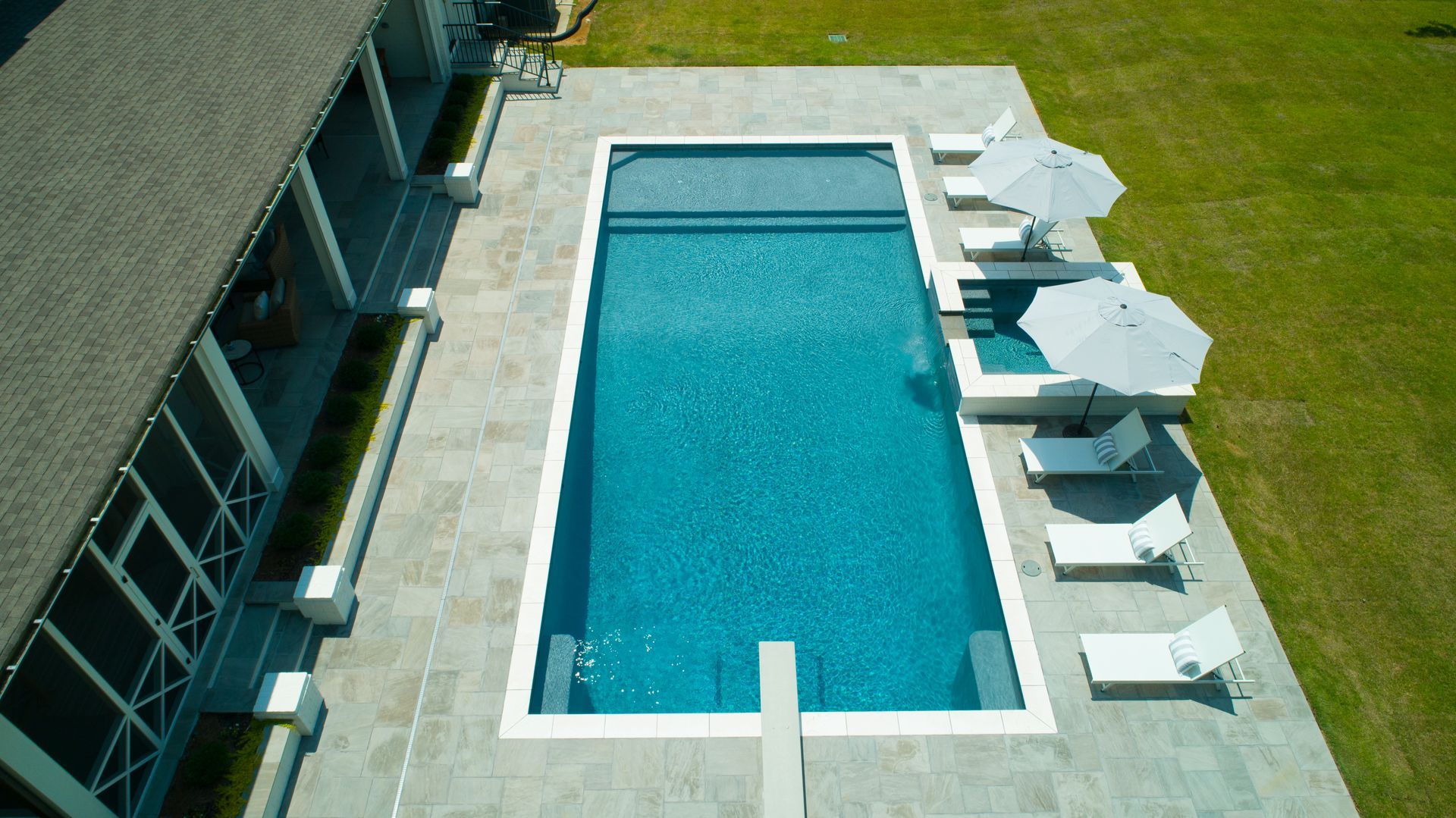 Aerial view of a rectangular turquoise pool with lounge chairs, umbrellas, and a house on the side.