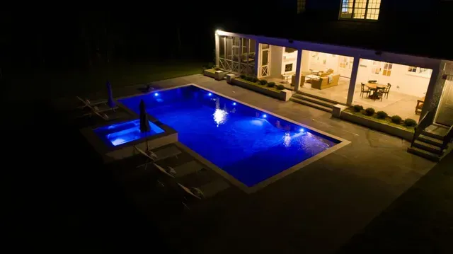 Night view of a pool with blue lights next to a house with lit windows, showing a dining area.