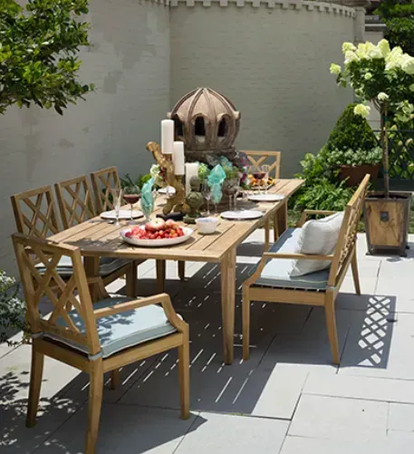 Outdoor dining table with chairs, set for a meal. Light wood furniture on a stone patio with plants and a decorative structure.