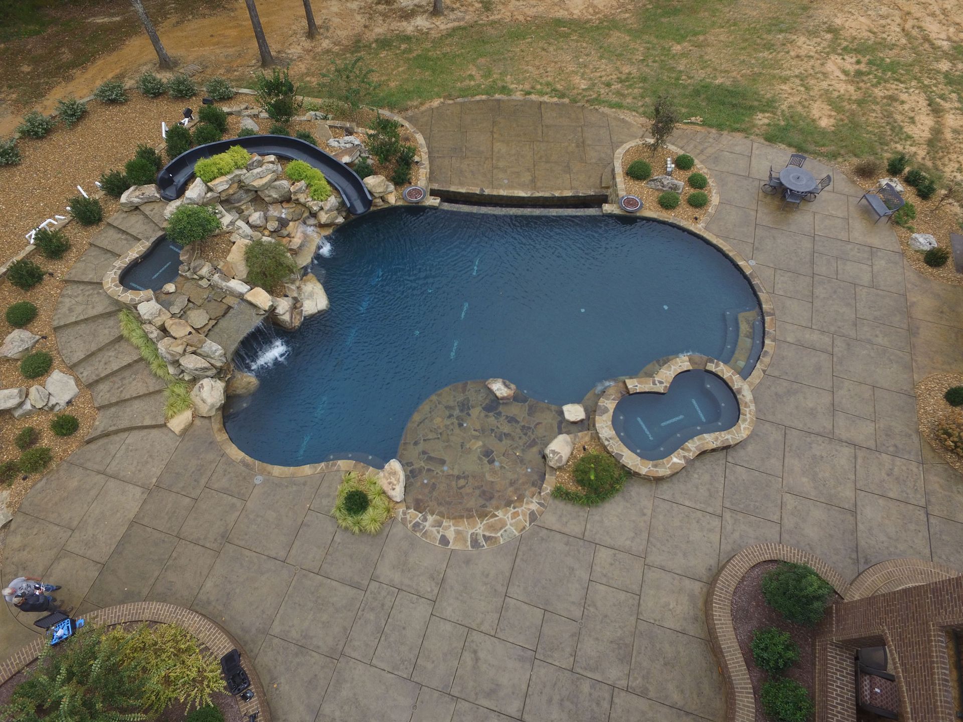 Aerial view of a dark blue, free-form swimming pool with a waterfall, spa, and stone patio.