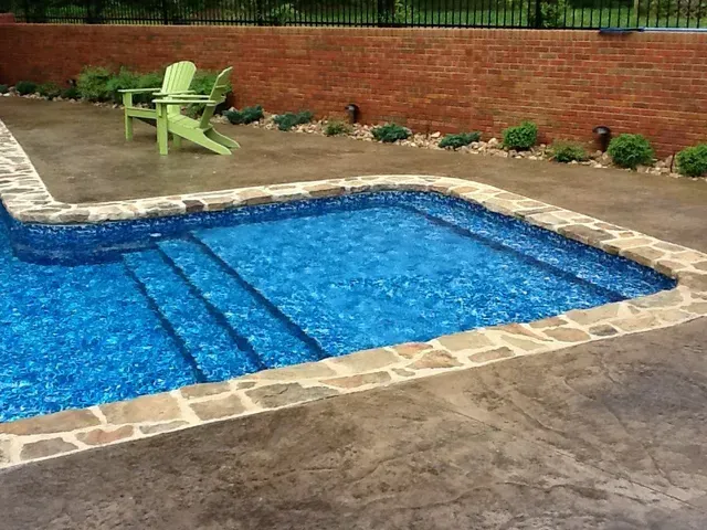 Swimming pool with stone border, steps, blue water. Two green chairs sit nearby a brick wall.