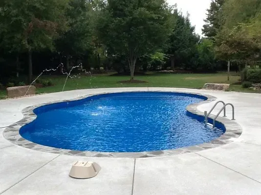 A backyard pool with blue water and a decorative fountain, surrounded by a concrete patio and green lawn.