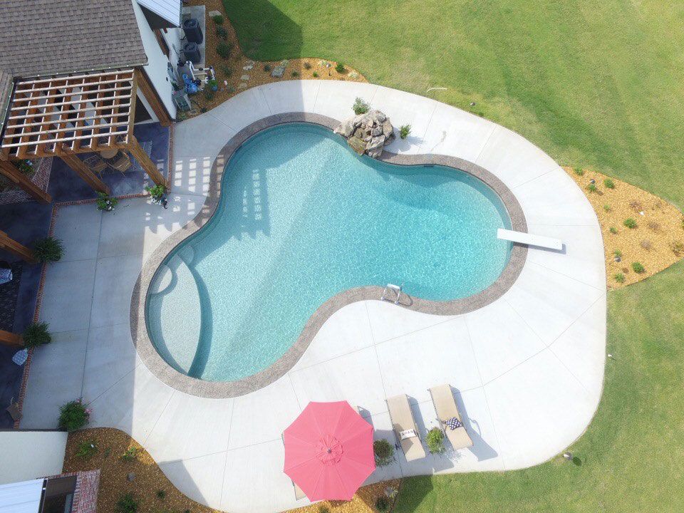 Aerial view of a freeform swimming pool with a diving board, surrounded by concrete and grass.