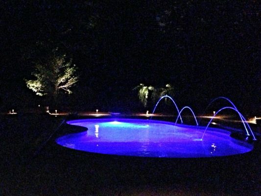Nighttime view of a pool with blue underwater lights and illuminated water fountains. Ambient lighting in the background.