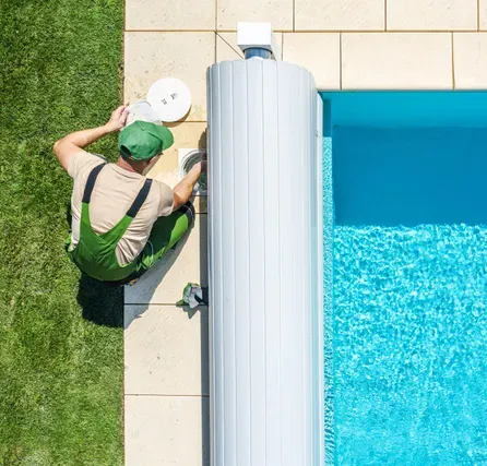 Person in green overalls working on a pool cover next to a pool, on a sunny day.