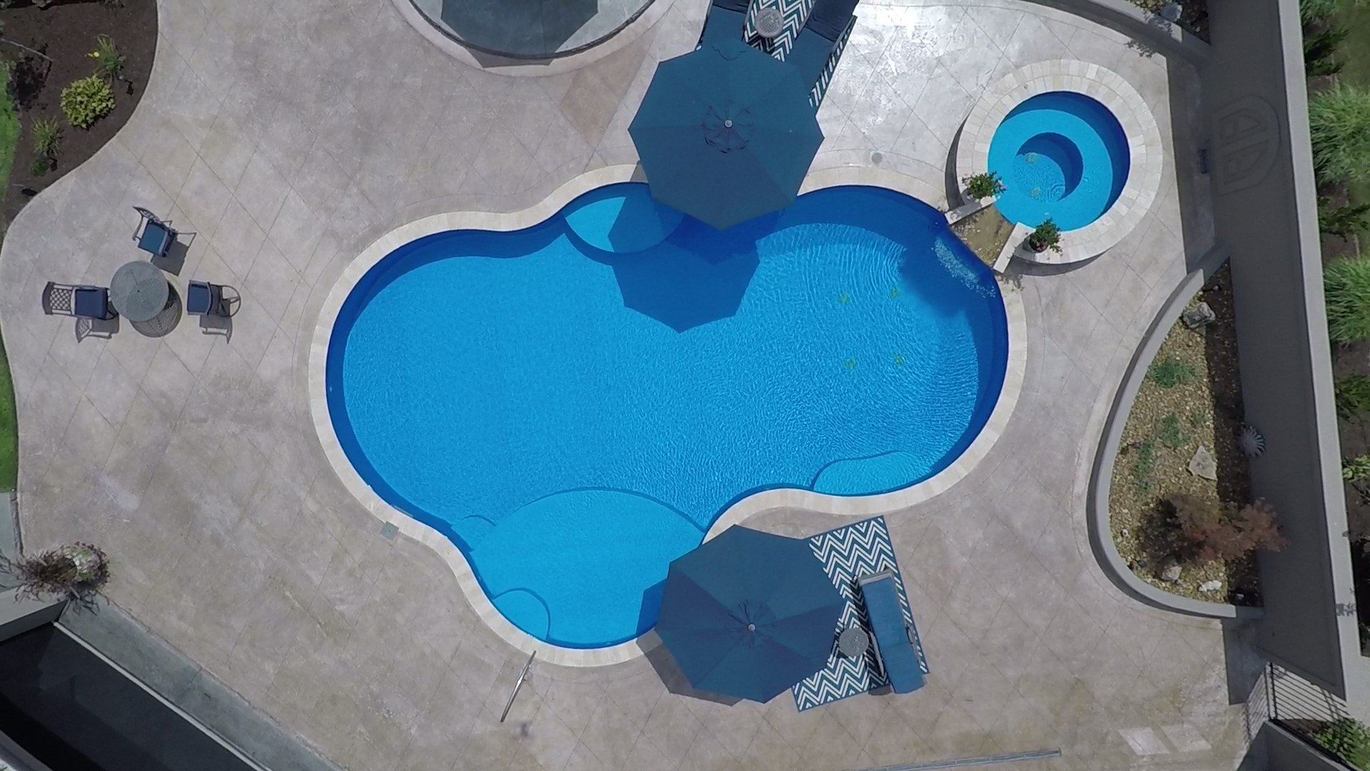 Overhead view of a blue freeform pool with a hot tub, surrounded by concrete decking, lounge chairs, and umbrellas.