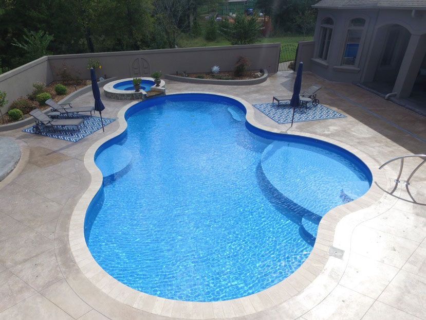 Blue, freeform swimming pool surrounded by a beige patio, with a spa and lounge chairs.