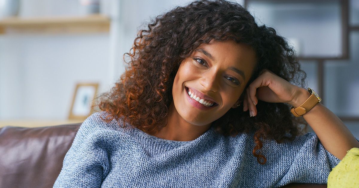 A woman is sitting on a couch with her hand on her head and smiling.