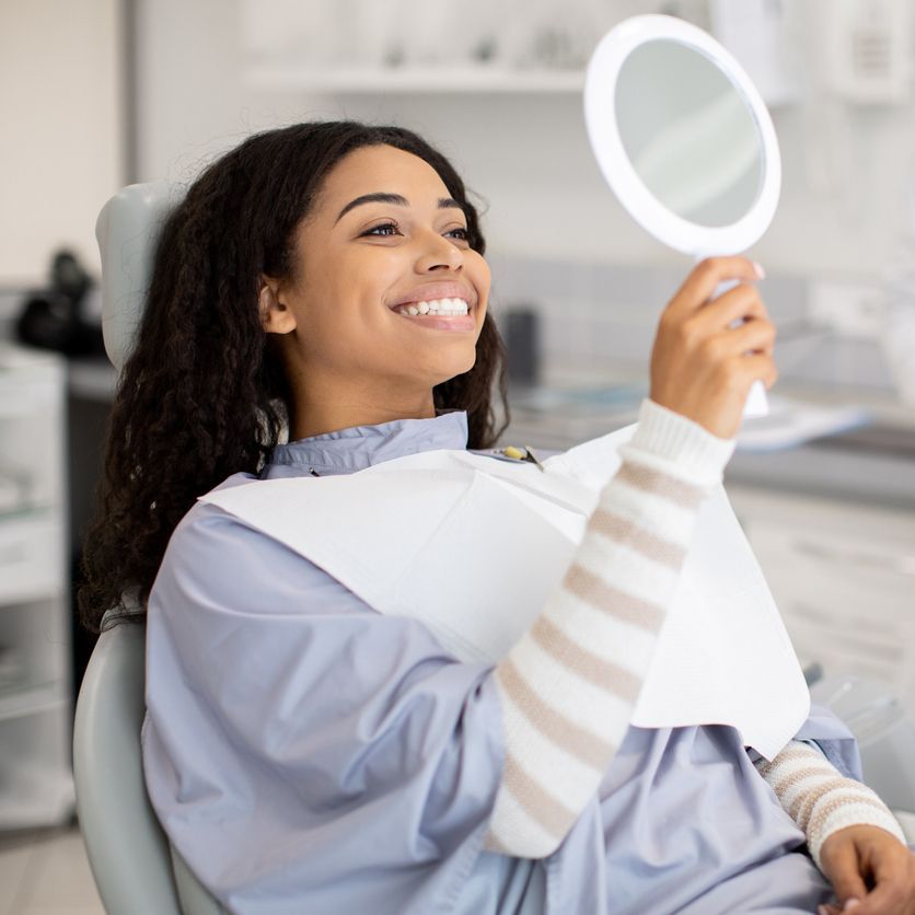 A woman is sitting in a dental chair holding a mirror and smiling.