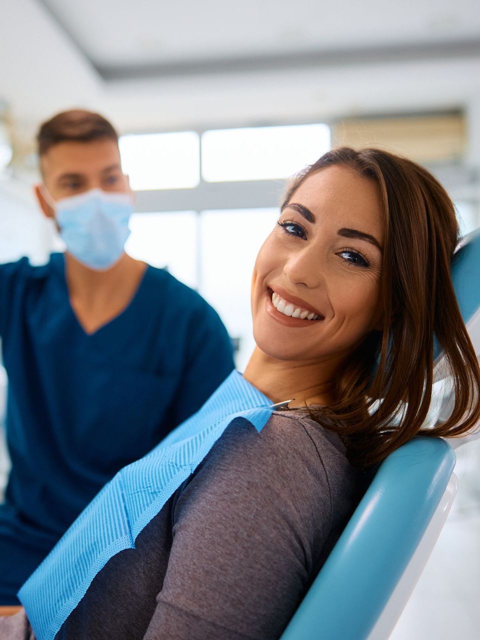 A woman is smiling while sitting in a dental chair next to a dentist wearing a mask.