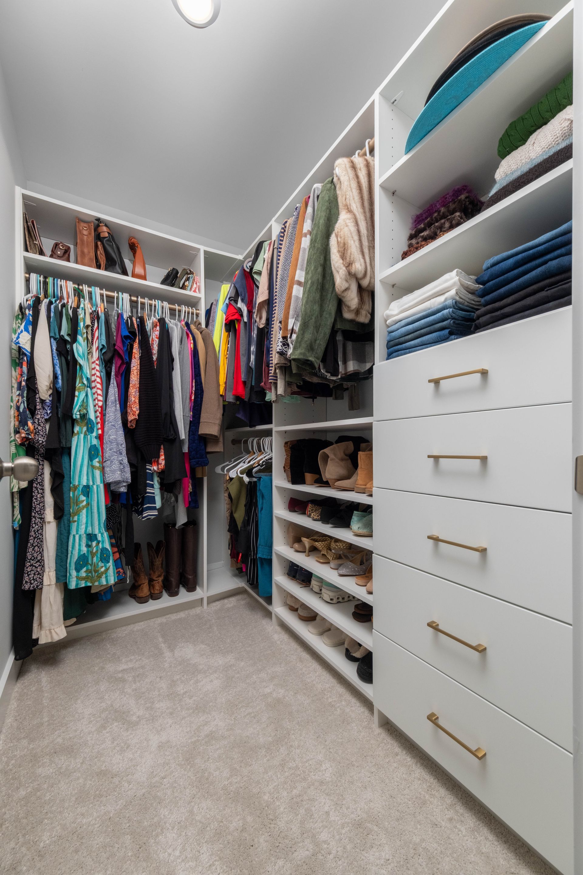 Walk-in closet with white shelves and drawers, filled with clothes and shoes. Gray carpet and neutral walls.