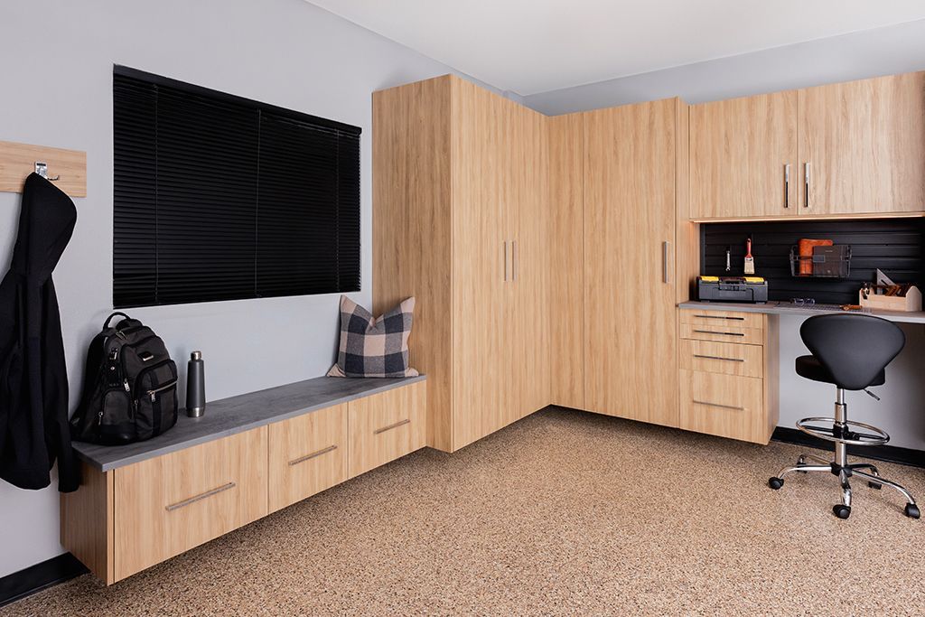 Modern office corner with wood cabinets, black desk, chair, backpack, and window bench.