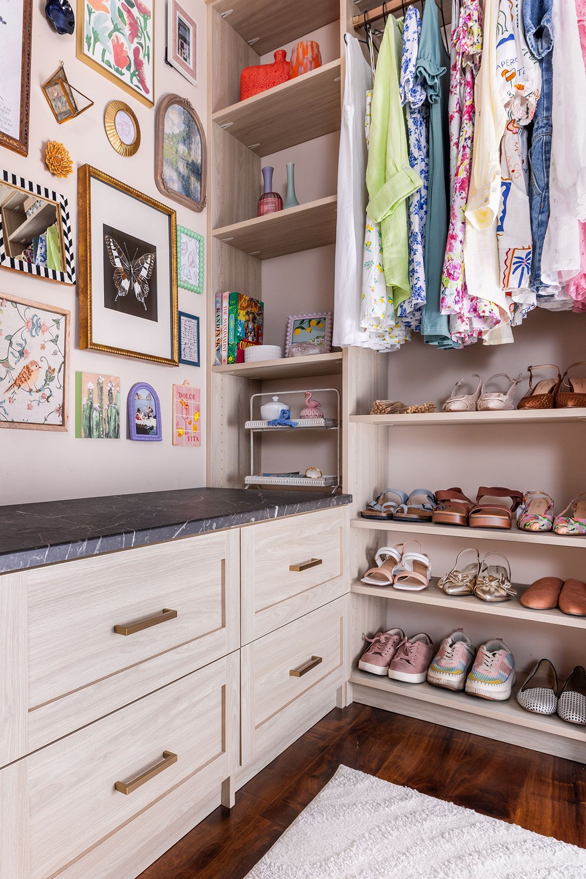 A walk-in closet with white drawers, hanging clothes, open shoe shelves, and framed wall art.