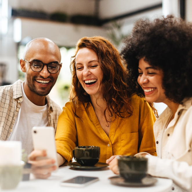 A group of people are sitting at a table laughing and looking at a cell phone.