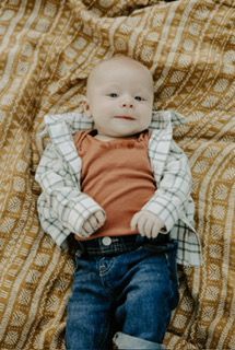 Baby lying on patterned blanket wearing a rust-colored shirt, jeans, and a plaid shirt.