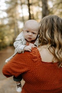 Baby looks over shoulder from person's shoulder, wearing plaid, fall foliage in background.