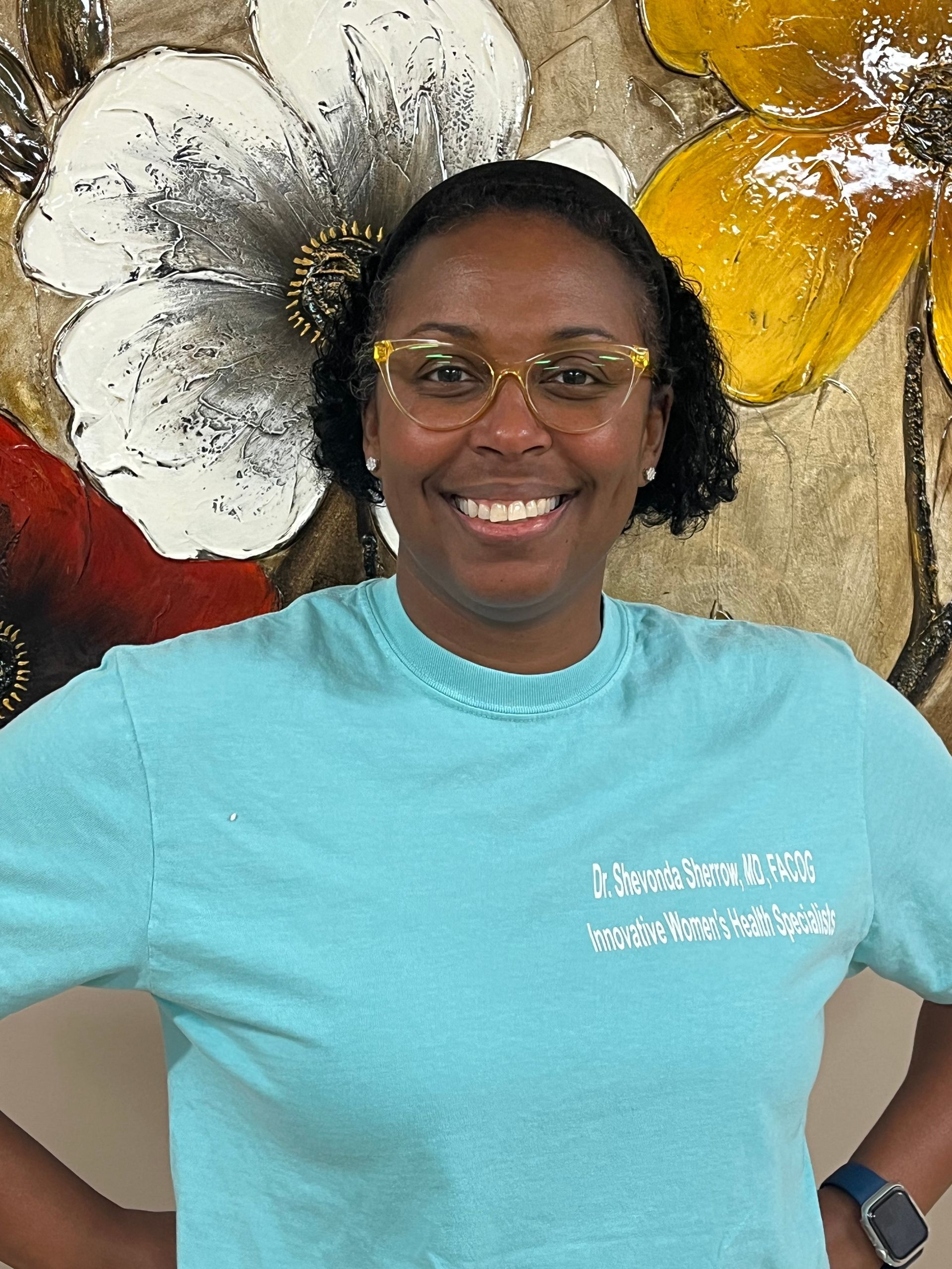 A woman wearing glasses and a blue shirt is smiling in front of a painting of flowers.
