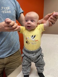 Baby in yellow onesie and gray pants is held up by an adult; orange background.