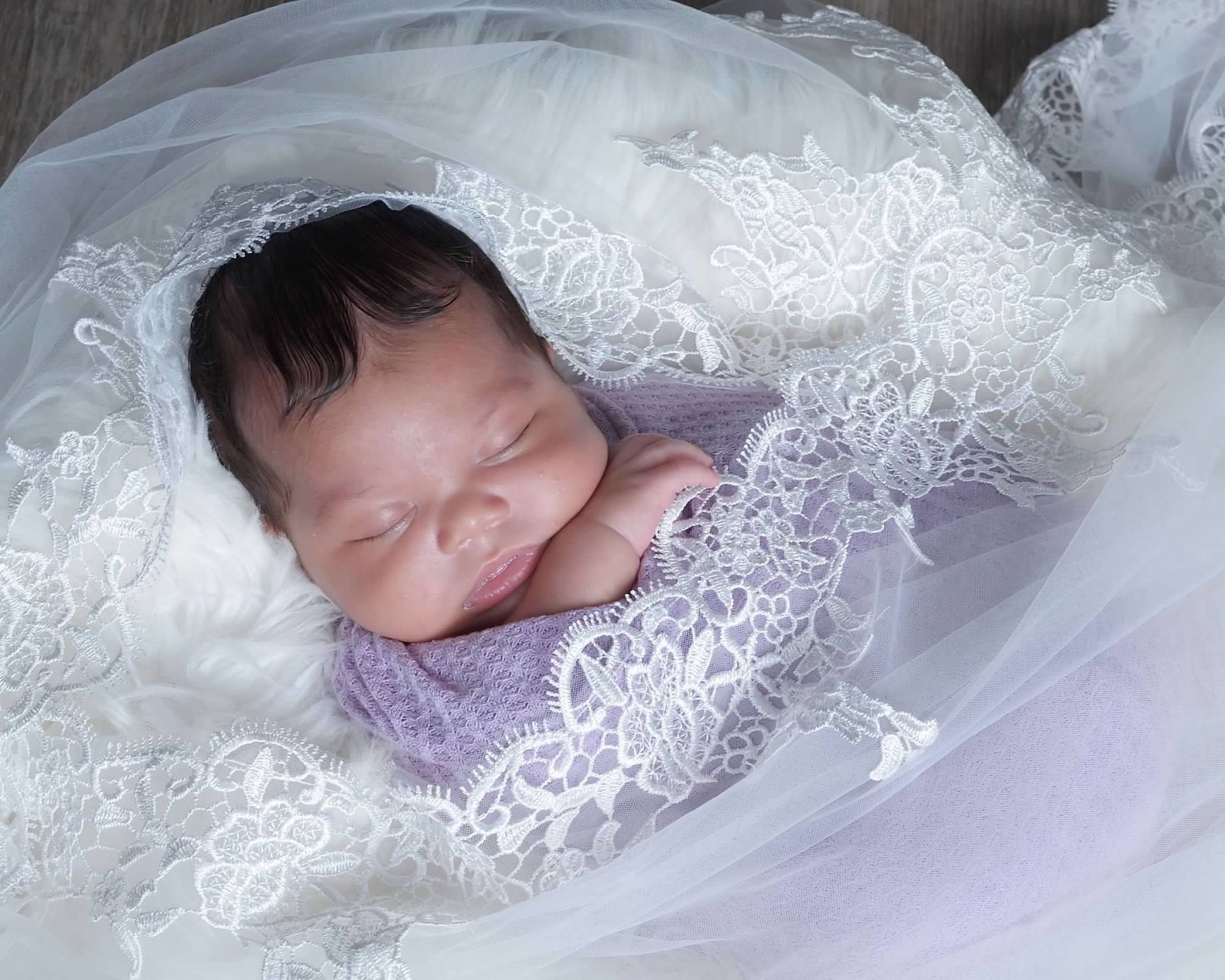 Newborn baby swaddled in purple blanket, nestled on white fur, surrounded by white lace and tulle.