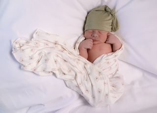 Newborn baby swaddled in a patterned blanket, wearing a green hat, resting on white sheets.