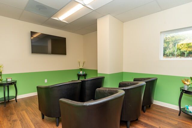 A waiting room with black armchairs, a TV, and small tables. Green and cream walls, a window.