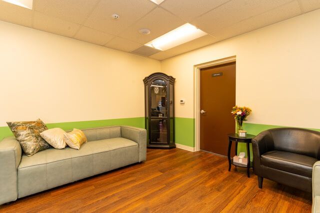 Waiting room with couch, armchair, cabinet, and door; green and cream walls, wood floor.