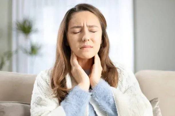 A woman holds her throat with one hand while gently rubbing it with the other, indicating discomfort from a sore throat.