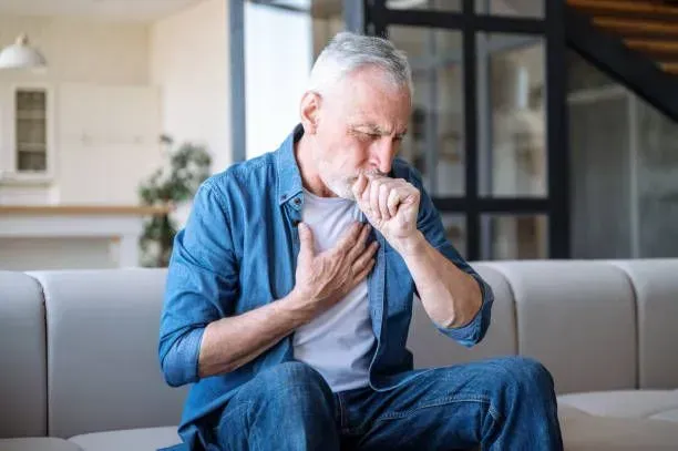 A man sits on a couch with his hands on his chest, appearing to cough or experience discomfort.