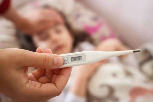 A caregiver holds an electronic thermometer in front of a child, monitoring symptoms of Hand Foot and Mouth Disease.