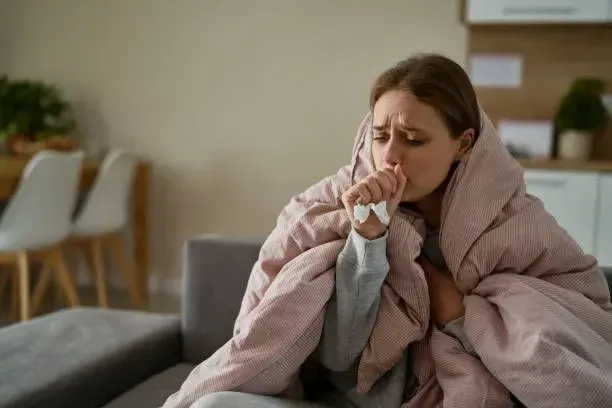 A woman sits on a couch, wrapped in a blanket, appearing cozy while dealing with a cough.