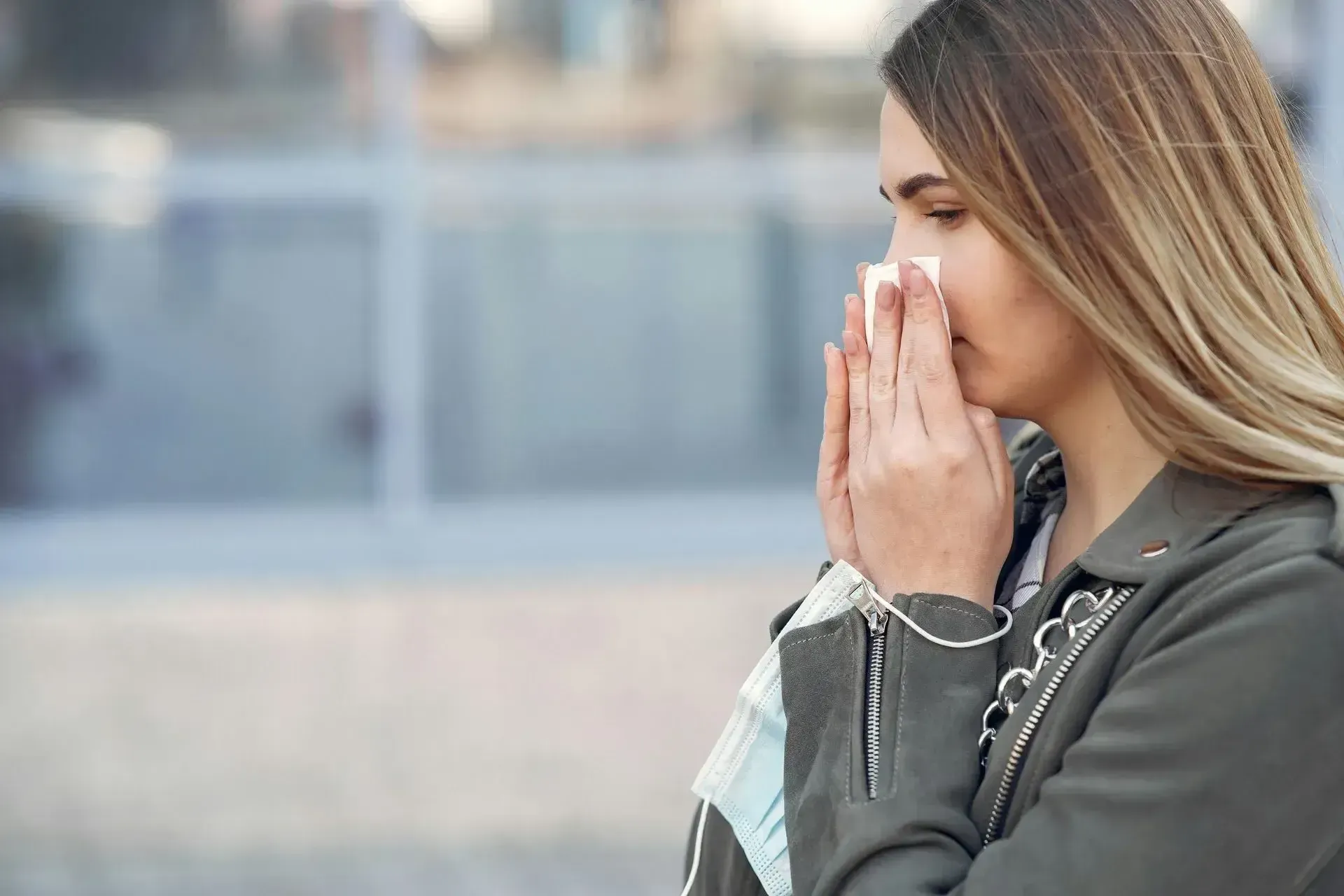 A woman outdoors is blowing her nose, visibly affected by allergies.