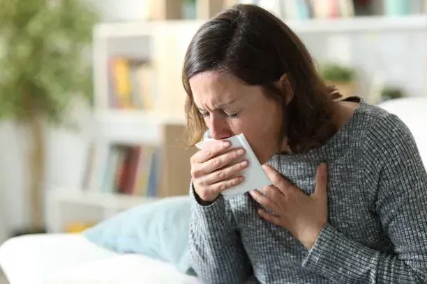 A woman sitting on a couch blows her nose, visibly affected by bronchitis symptoms.